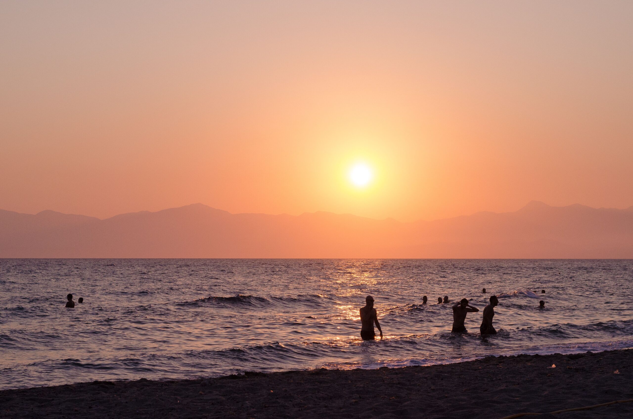 A beautiful sunset over a Greek beach with silhouettes of people swimming and playing in the sea.